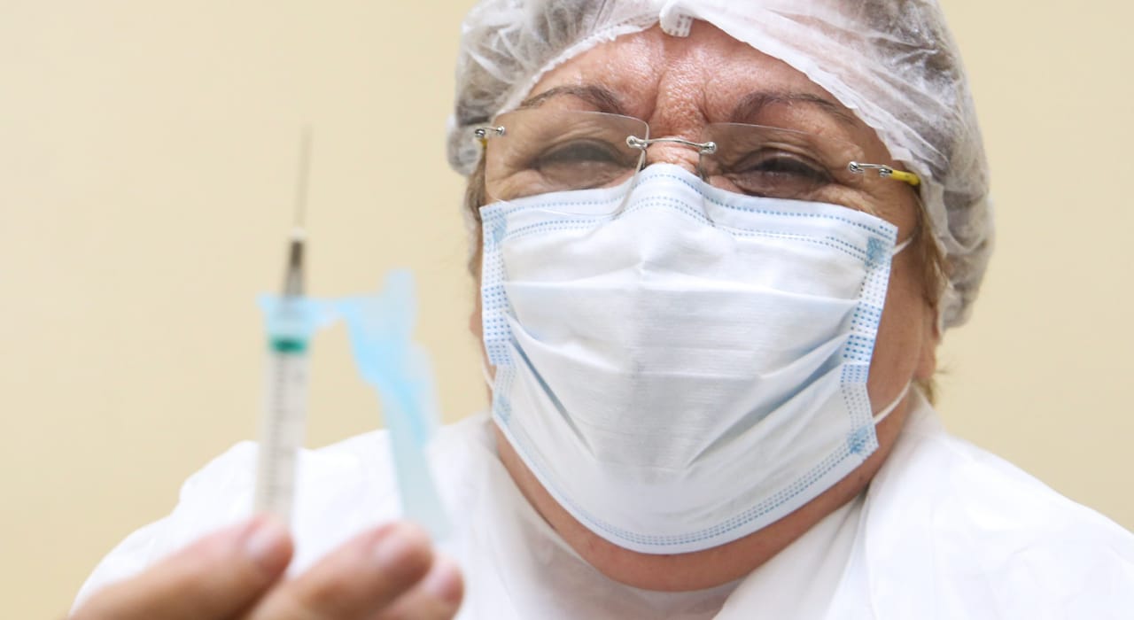 enfermeira socorro bezerra posa para a foto segurando uma seringa, apesar de ela estar com máscara, dá para perceber que ela está sorrindo